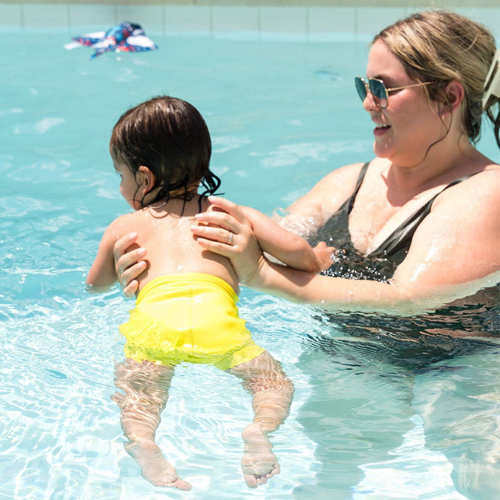 Little one swimming with mum in the pool, wearing neon yellow swim diaper.