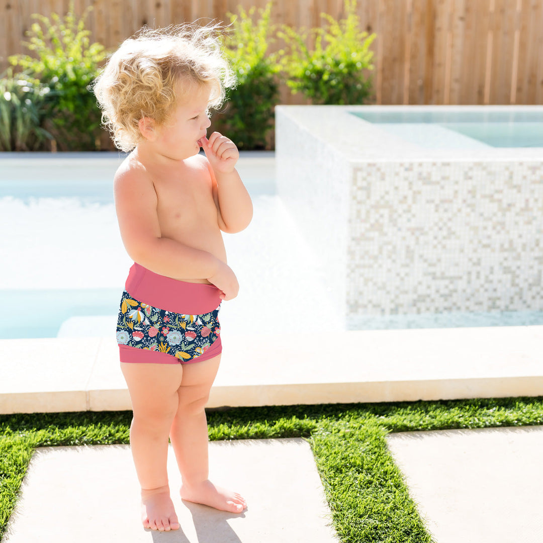 Little one wearing neoprene swim diaper in navy with pink trims, and floral themed print.