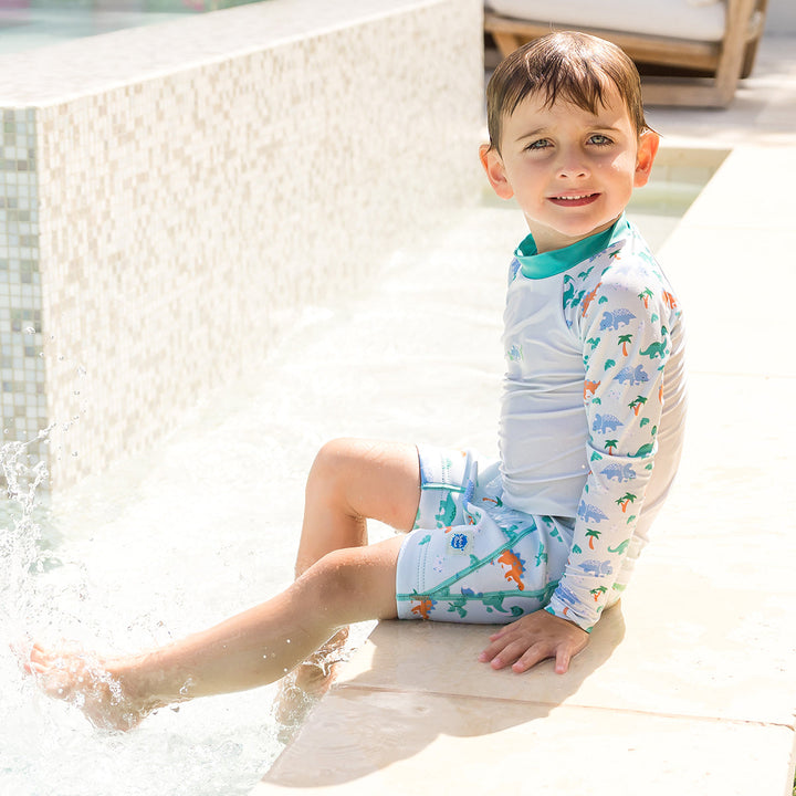 Child wearing neoprene swim shorts in mint and dinosaurs themed print. He's also wearing matching rash top.