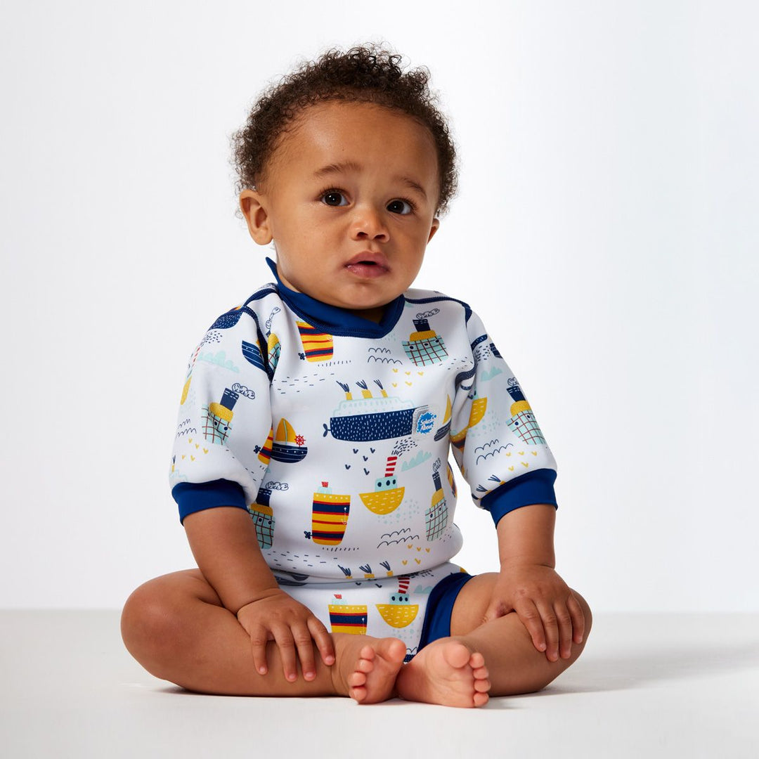 Lifestyle image of child wearing a baby wetsuit with built in swim diaper in white with navy trims and boats themed print. Front.