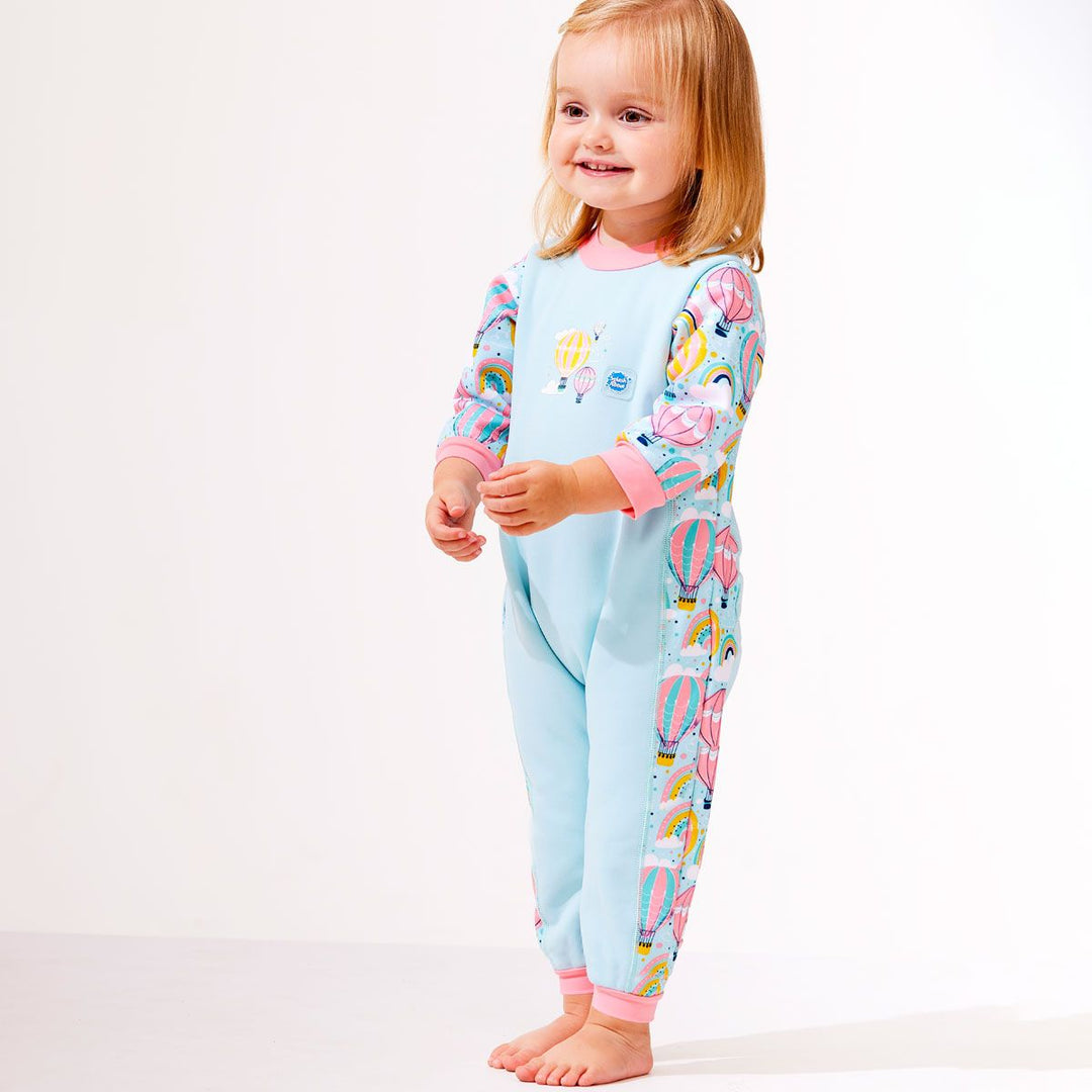 Lifestyle image of toddler wearing a fleece-lined baby wetsuit in baby blue with pink trims and hot air balloons themed print, including rainbows and clouds. 